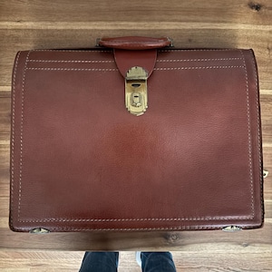 May include: A rectangular brown leather briefcase featuring a top handle and a central gold-toned metal latch closure. The exterior displays visible stitching along the edges and two small metal accents at the bottom corners of the front panel.