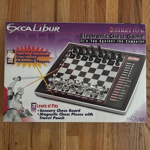 Peut inclure: Boîte de jeu d'échecs électronique Excalibur Stiletto II. La boîte présente un échiquier avec des pièces noires et argentées. Le texte sur la boîte comprend "Electronic Chess Game" et "It's You Against the Computer."