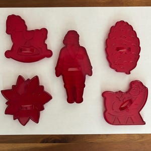 May include: A set of five red plastic cookie cutters featuring holiday-themed shapes including a rocking horse, a standing Santa Claus figure, a pinecone, a poinsettia flower, and a Santa Claus sitting in a sleigh, all arranged on a white surface.