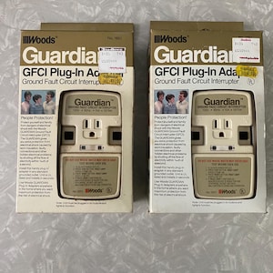 May include: Two white boxes with gold trim, each containing a Woods Guardian GFCI Plug-In Adapter. The boxes have a picture of a white electrical outlet with a test button and a reset button. The text on the boxes reads "Guardian Ground Fault Circuit Interrupter".