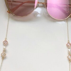 May include: A pair of pink sunglasses with a gold chain attached to each arm. The chain has a pink and white clover charm with a pearl bead.