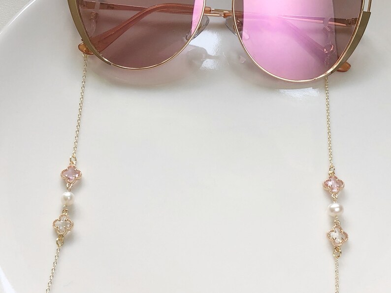 May include: A pair of pink sunglasses with a gold chain attached to each arm. The chain has a pink and white clover charm with a pearl bead.