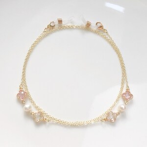 May include: A gold chain with pink and white flower charms and pearl beads. The chain has two loops at the ends for attaching to glasses.