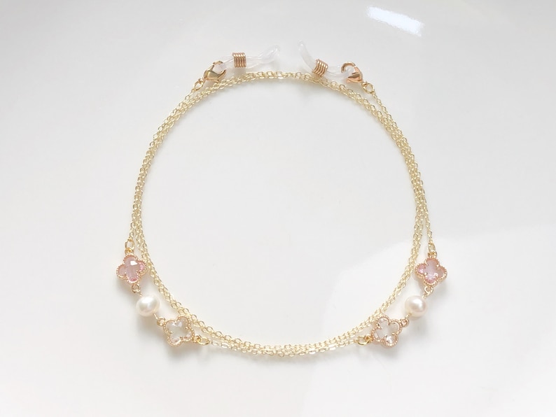 May include: A gold chain with pink and white flower charms and pearl beads. The chain has two loops at the ends for attaching to glasses.