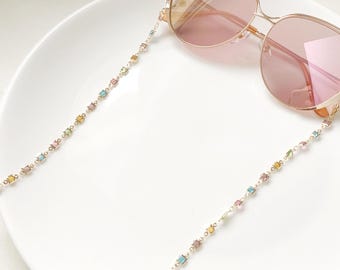 May include: Gold-toned sunglasses with pink lenses and a delicate chain. The chain is adorned with small, colorful, square-shaped beads in pastel hues. The glasses rest on a white surface, showcasing the accessory's design.