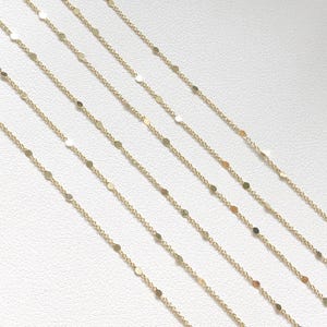 Op de afbeelding: Meerdere gouden kettingen met kleine, ronde, platte, gelijkmatig verdeelde schakels. De kettingen zijn waaiervormig op een witte achtergrond gerangschikt.
