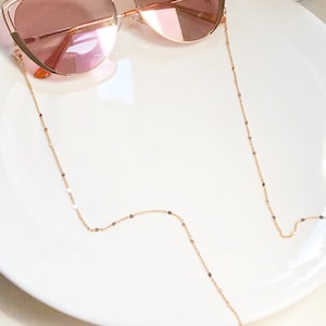 Op de afbeelding: Een gouden ketting met kleine, ronde schakels, ontworpen om een zonnebril te houden. De ketting ligt op een wit bord.