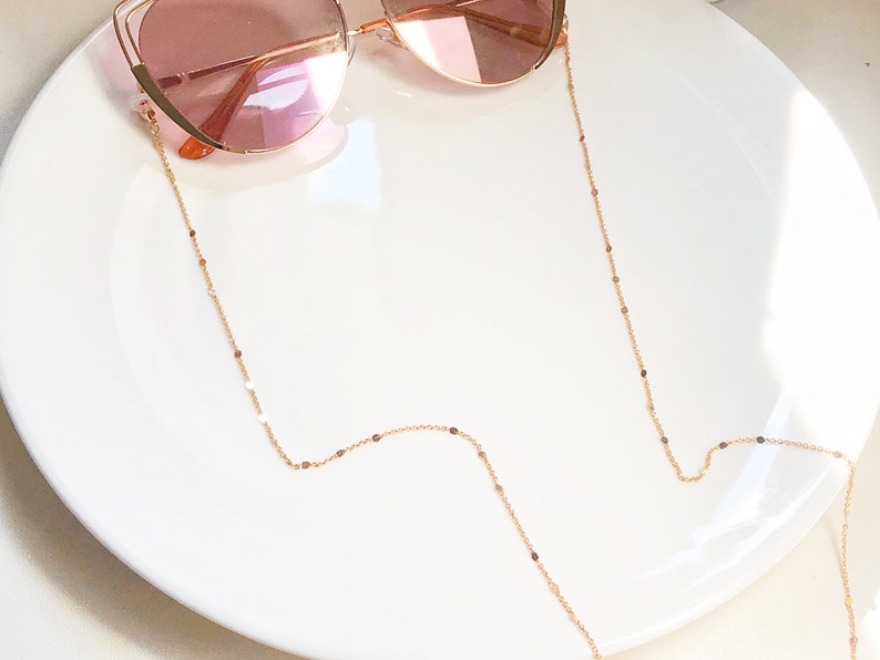 Op de afbeelding: Een gouden ketting met kleine, ronde schakels, ontworpen om een zonnebril te houden. De ketting ligt op een wit bord.