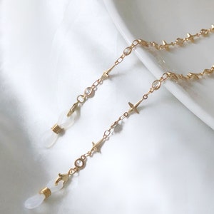 Könnte beinhalten: Goldfarbene Brillenketten mit Stern- und Kristallakzenten. Die Kette hat kleine sternförmige Anhänger und klare Kristalle mit Schlaufen zum Befestigen an Brillen. Die Kette wird auf einer weißen Oberfläche präsentiert.