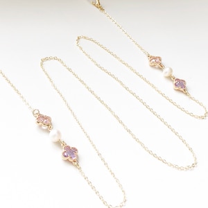 May include: A gold chain with two pink and purple flower charms and a pearl bead on each side. The chain is designed to hold glasses.