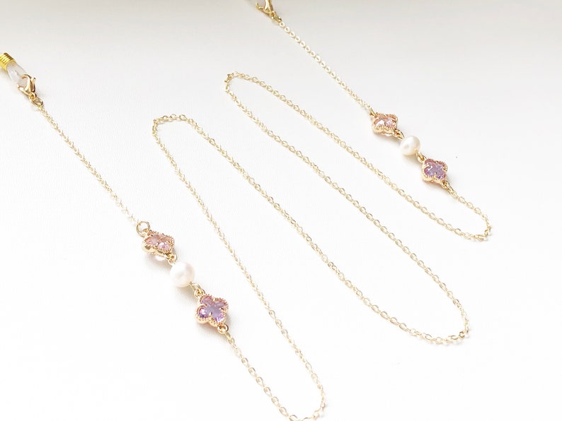 May include: A gold chain with two pink and purple flower charms and a pearl bead on each side. The chain is designed to hold glasses.