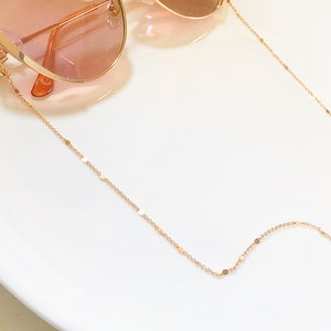 Op de afbeelding: Een gouden ketting met kleine, platte, ronde schakels. De ketting is ontworpen om brillen vast te houden.