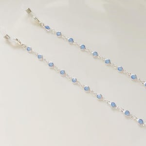Op de afbeelding: Een paar zilveren brillenkettingen met lichtblauwe kralen. De kettingen zijn bevestigd aan transparante plastic oorstukken.