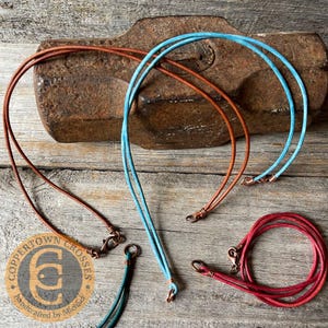 May include: Leather necklaces in various colors, including brown, blue, and red, with copper clasps. The necklaces are displayed on a wooden surface with a rusty metal object in the background. The logo for Coppertown Crosses is also visible.