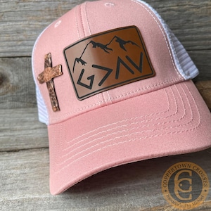 May include: A pink baseball cap with a white mesh back and a curved brim. The cap features a brown leather patch with the text "G>AV" and mountain silhouettes. A copper cross is attached to the side.