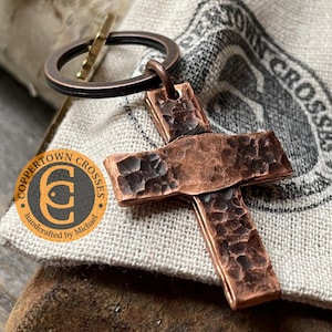 May include: A hammered copper cross keychain with a textured finish. The keychain is attached to a brown metal ring. The cross is on a white fabric background with a brown logo that reads "Coppertown Crosses handcrafted by Michael".