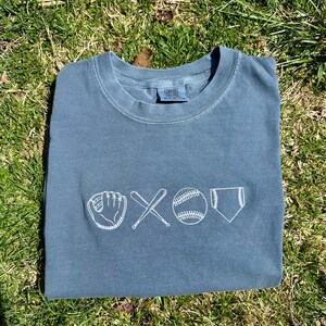 May include: A blue t-shirt with a white embroidered design of a baseball glove, two crossed bats, a baseball, and home plate.