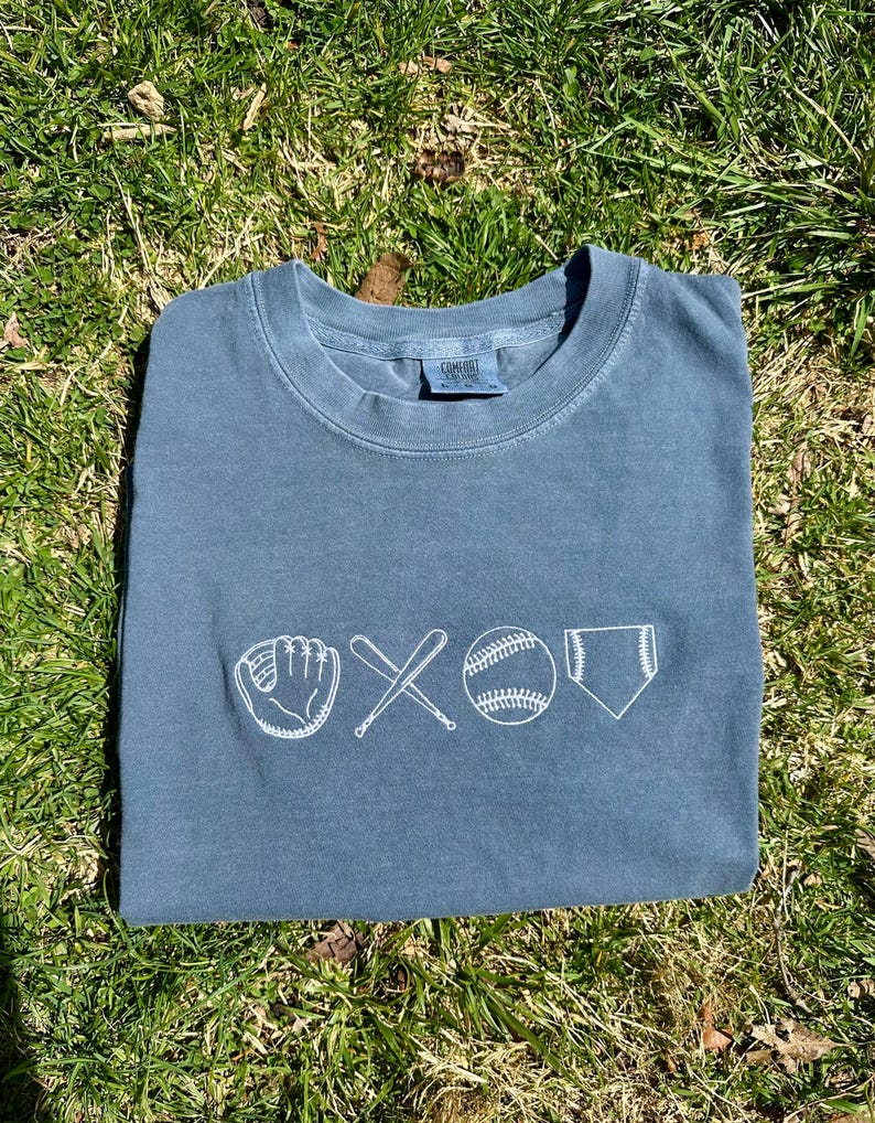 May include: A blue t-shirt with a white embroidered design of a baseball glove, two crossed bats, a baseball, and home plate.