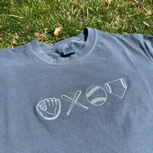 May include: A grey t-shirt with a white embroidered design of a baseball glove, two crossed bats, a baseball, and a home plate.