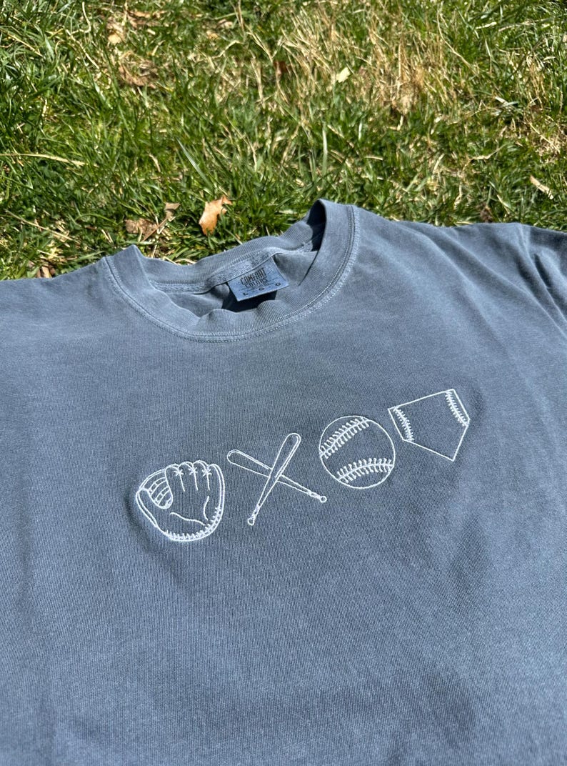 May include: A grey t-shirt with a white embroidered design of a baseball glove, two crossed bats, a baseball, and a home plate.