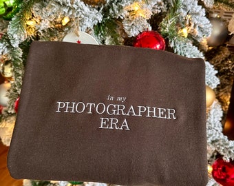 May include: A brown sweatshirt with the text "in my PHOTOGRAPHER ERA" embroidered in white.