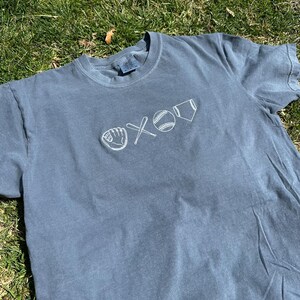 May include: A blue t-shirt with a white embroidered design of a baseball, bat, glove, and home plate.