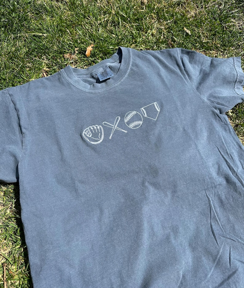 May include: A blue t-shirt with a white embroidered design of a baseball, bat, glove, and home plate.