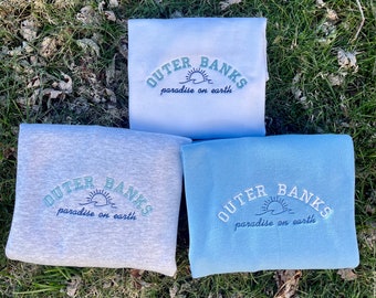 Puede incluir: Tres sudaderas con el texto "Outer Banks paradise on earth" en una fuente arqueada azul y blanca. Las sudaderas son de color gris, blanco y azul claro.