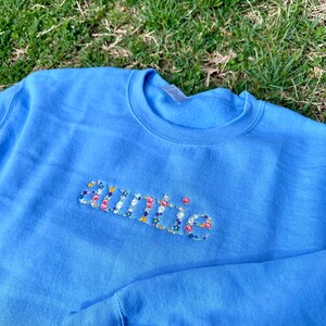 K&ouml;nnte beinhalten: Ein hellblauer Pullover mit dem Wort "auntie", das mit bunten Blumen bestickt ist.