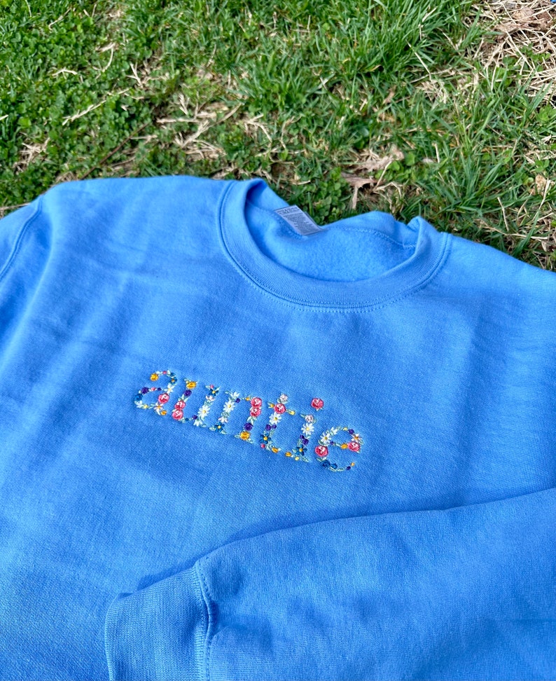 K&ouml;nnte beinhalten: Ein hellblauer Pullover mit dem Wort "auntie", das mit bunten Blumen bestickt ist.