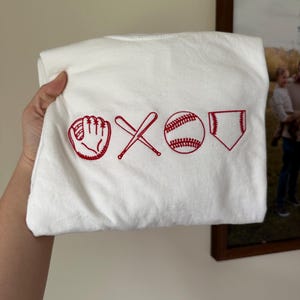 May include: White t-shirt with red embroidered baseball icons: a glove, two crossed bats, a baseball, and a home plate.