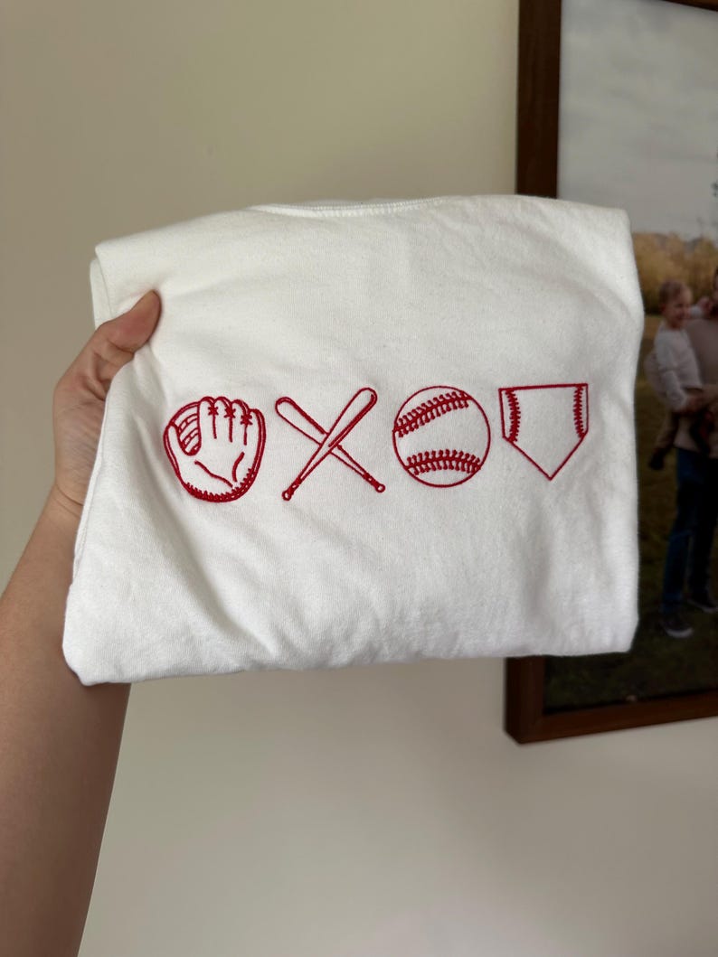 May include: White t-shirt with red embroidered baseball icons: a glove, two crossed bats, a baseball, and a home plate.