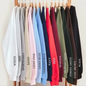 May include: A row of 14 sweatshirts hanging on a rack, each in a different color. The colors are white, ash, sports grey, sand, light pink, light blue, Carolina blue, navy, red, maroon, military green, forest green, dark brown, and black.