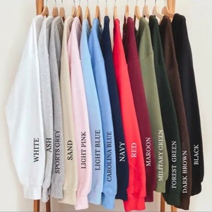 K&ouml;nnte beinhalten: Eine Reihe von 14 Sweatshirts, die an Kleiderb&uuml;geln h&auml;ngen und eine Vielzahl von Farben zeigen, darunter Wei&szlig;, Asche, Sportgrau, Sand, Hellrosa, Hellblau, Carolina-Blau, Marineblau, Rot, Weinrot, Milit&auml;rgr&uuml;n, Waldgr&uuml;n, Dunkelbraun und Schwarz. Jedes Sweatshirt hat den Farbnamen darunter gedruckt.