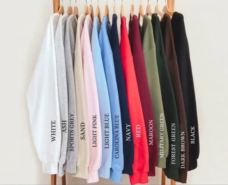 K&ouml;nnte beinhalten: Eine Reihe von 14 Sweatshirts, die an Kleiderb&uuml;geln h&auml;ngen und eine Vielzahl von Farben zeigen, darunter Wei&szlig;, Asche, Sportgrau, Sand, Hellrosa, Hellblau, Carolina-Blau, Marineblau, Rot, Weinrot, Milit&auml;rgr&uuml;n, Waldgr&uuml;n, Dunkelbraun und Schwarz. Jedes Sweatshirt hat den Farbnamen darunter gedruckt.
