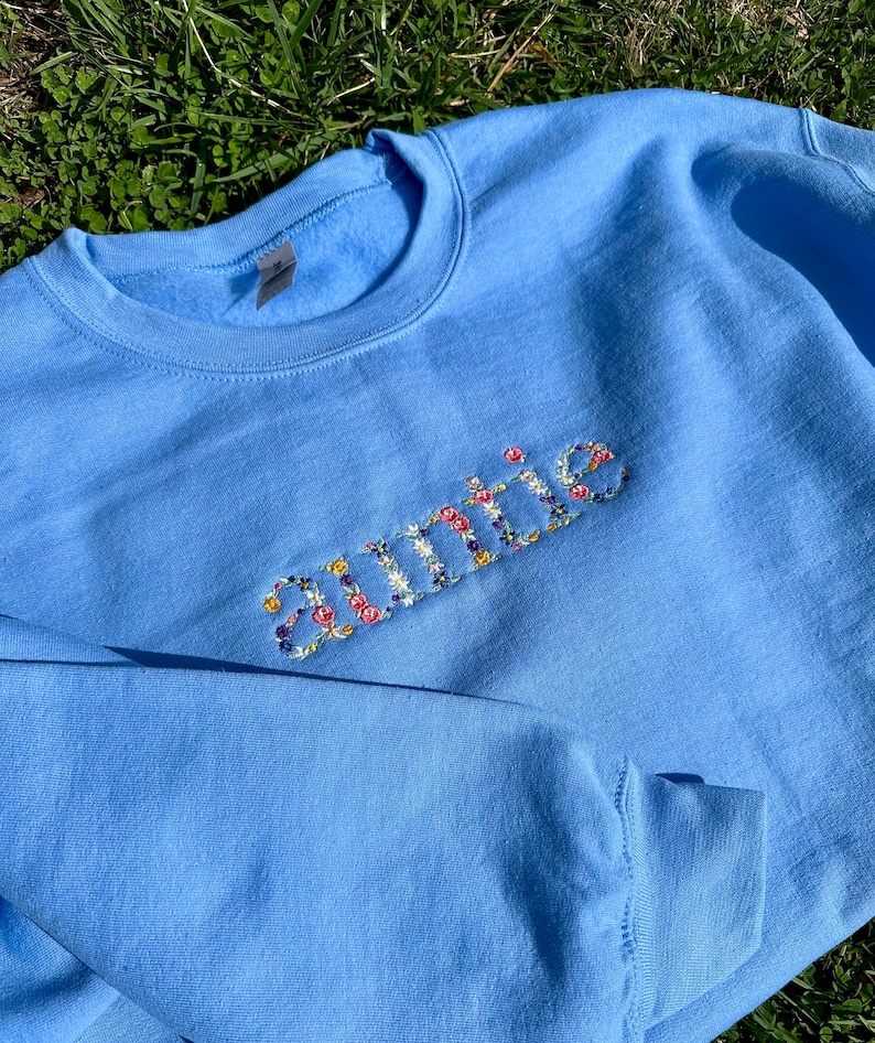 K&ouml;nnte beinhalten: Ein hellblauer Pullover mit dem Wort "Tante" in bunten Blumenbuchstaben gestickt.