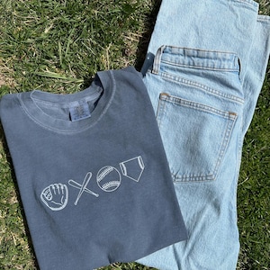 May include: A dark blue t-shirt with a white baseball-themed graphic. The graphic includes a baseball, bat, glove, and home plate. The t-shirt is folded and lying on top of a pair of light blue jeans.