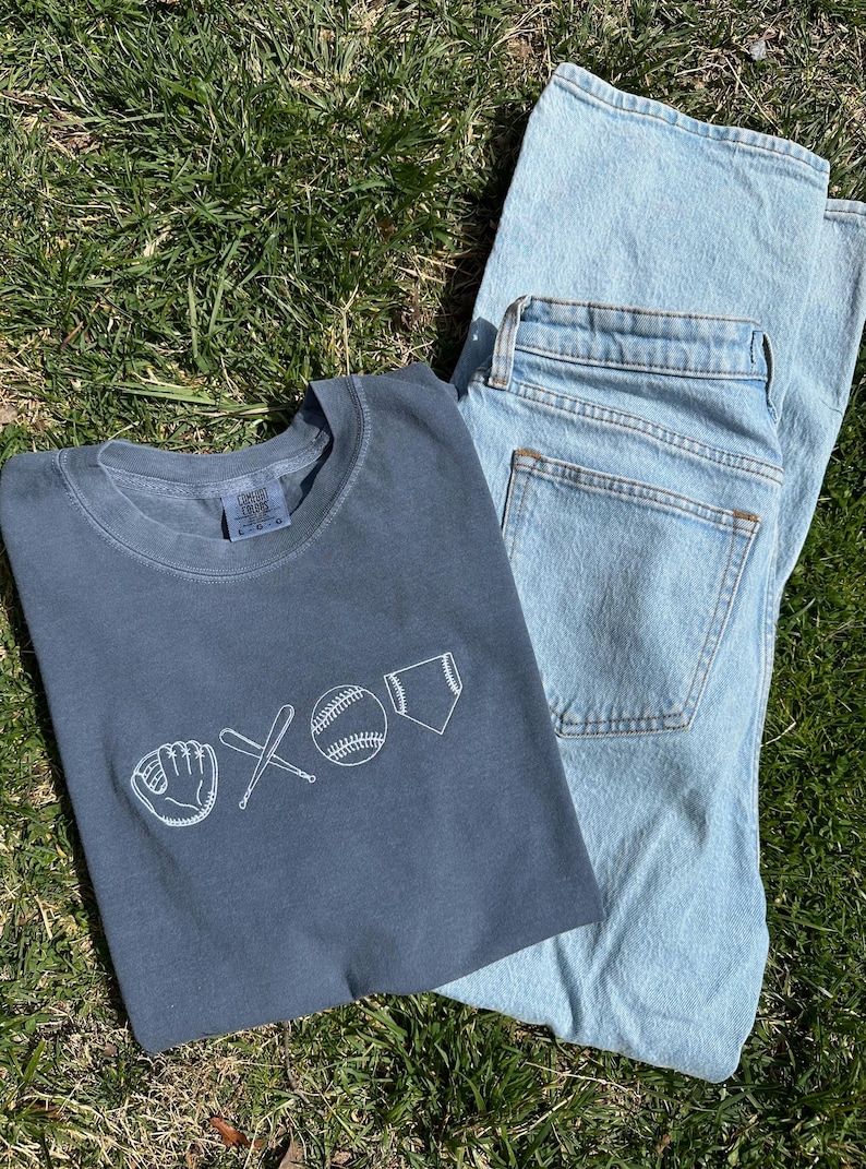 May include: A dark blue t-shirt with a white baseball-themed graphic. The graphic includes a baseball, bat, glove, and home plate. The t-shirt is folded and lying on top of a pair of light blue jeans.