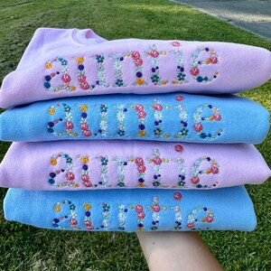 K&ouml;nnte beinhalten: Vier hellblaue und rosafarbene Sweatshirts, die &uuml;bereinander gestapelt sind. Jedes Sweatshirt hat das Wort "Auntie" in Wei&szlig; mit bunten Blumen gestickt.
