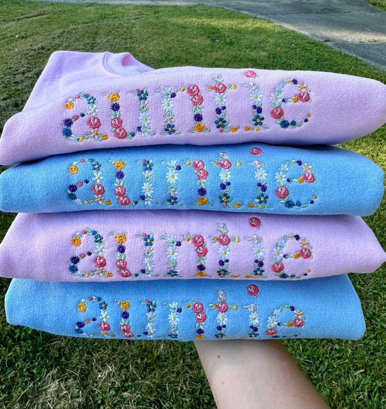 K&ouml;nnte beinhalten: Vier hellblaue und rosafarbene Sweatshirts, die &uuml;bereinander gestapelt sind. Jedes Sweatshirt hat das Wort "Auntie" in Wei&szlig; mit bunten Blumen gestickt.