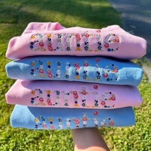 K&ouml;nnte beinhalten: Vier rosa und blaue Sweatshirts, die &uuml;bereinander gestapelt sind. Jedes Sweatshirt hat das Wort "Tante" in bunten Blumen gestickt.