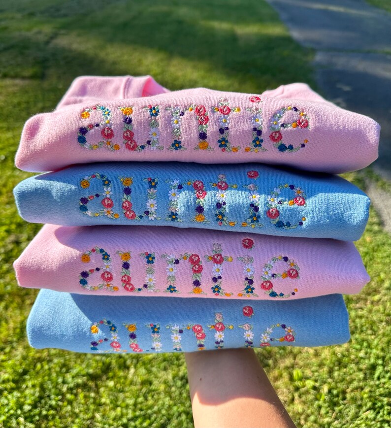 K&ouml;nnte beinhalten: Vier rosa und blaue Sweatshirts, die &uuml;bereinander gestapelt sind. Jedes Sweatshirt hat das Wort "Tante" in bunten Blumen gestickt.