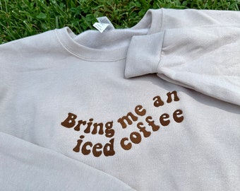 May include: A light beige sweatshirt with brown embroidered text that reads "Bring me an iced coffee".