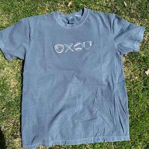 May include: A blue t-shirt with a white embroidered baseball design. The design includes a baseball glove, two bats crossed, a baseball, and home plate.