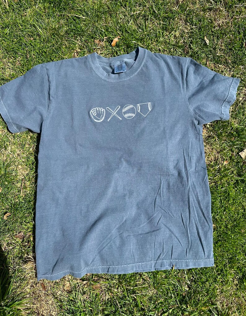 May include: A blue t-shirt with a white embroidered baseball design. The design includes a baseball glove, two bats crossed, a baseball, and home plate.