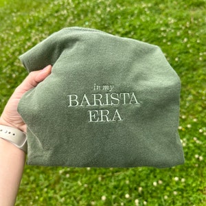 May include: A green t-shirt with the text "In my Barista Era" embroidered in white.