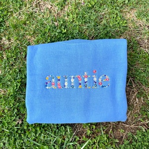 K&ouml;nnte beinhalten: Ein hellblauer Pullover mit dem Wort "auntie" in bunten Blumen gestickt.