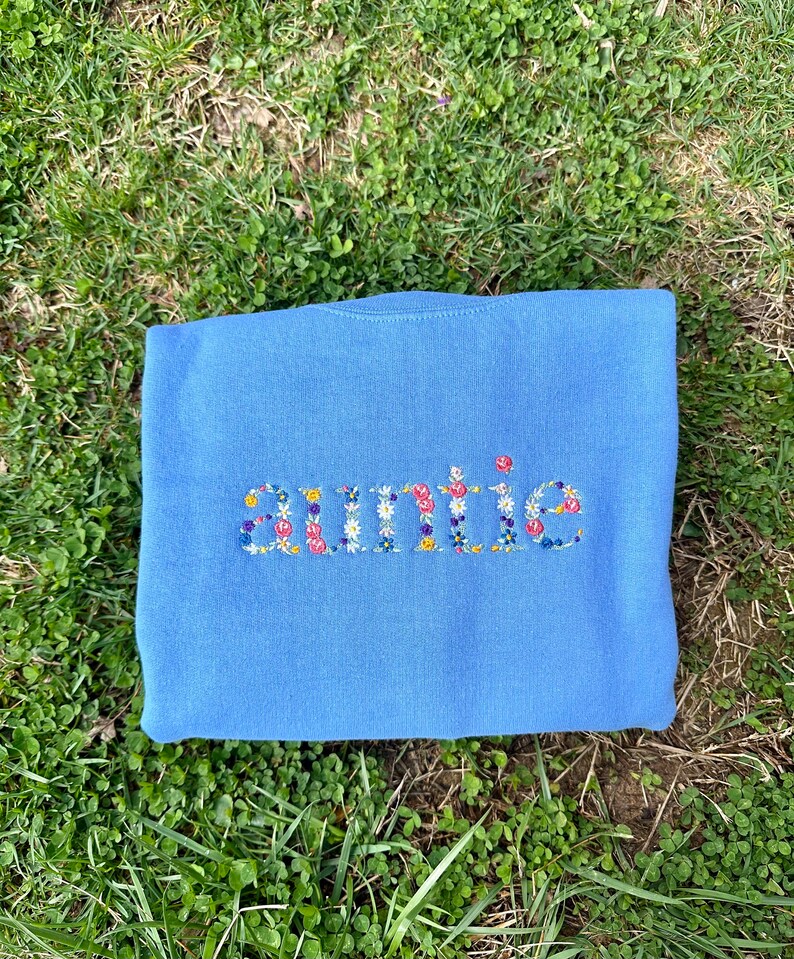 K&ouml;nnte beinhalten: Ein hellblauer Pullover mit dem Wort "auntie" in bunten Blumen gestickt.