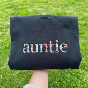 K&ouml;nnte beinhalten: Schwarzer Pullover mit dem Wort "auntie" in bunten Blumen gestickt.