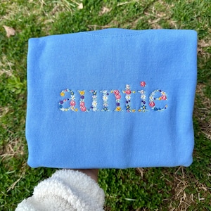 K&ouml;nnte beinhalten: Ein hellblauer Pullover mit dem Wort "auntie", das mit bunten Blumen bestickt ist.